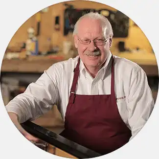 Eric Lindeblad, a senior craftsman with an apron, stands in a woodworking shop, representing the third generation of Lindeblad Piano Restoration.