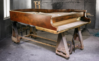 Wood-tone piano on sawhorses in a refinishing booth.