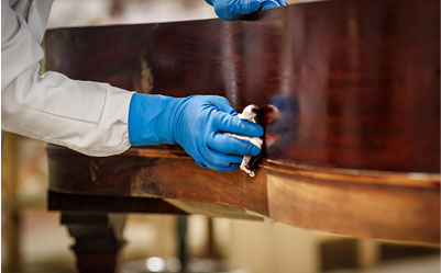 Hand in blue rubber glove applying dark wood stain to a piano case.